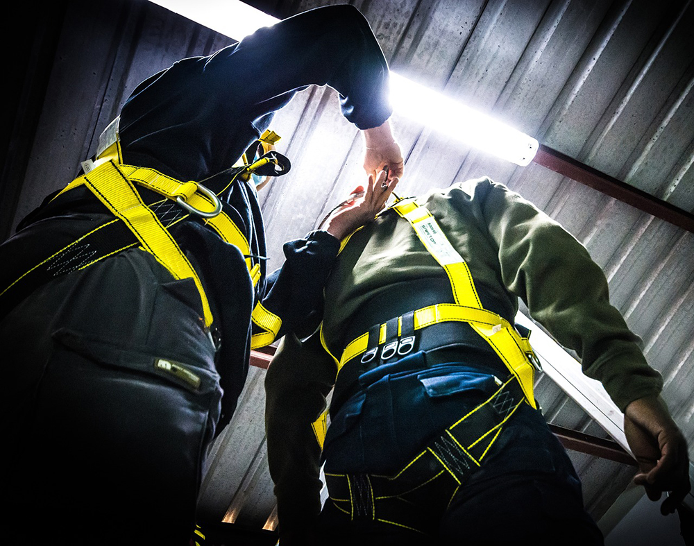 two workers wearing safety harnesses for work at height. One person is inspecting the other's harness