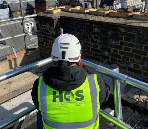 Person inspecting a guardrail on a roof. He is wearing a hi-vis with HOS Fall Protection Specialists 