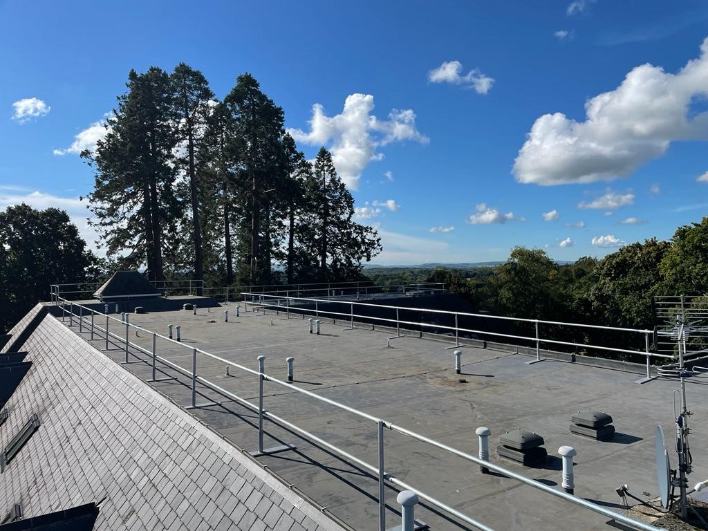 Flat roof with compliant metal fall protection guardrails installed around the perimeter, overlooking trees and countryside under a clear sky.