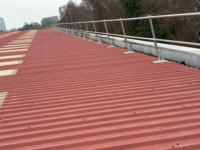 Long industrial metal roof with fixed fall protection guardrail installed along the edge to ensure safe working at height.